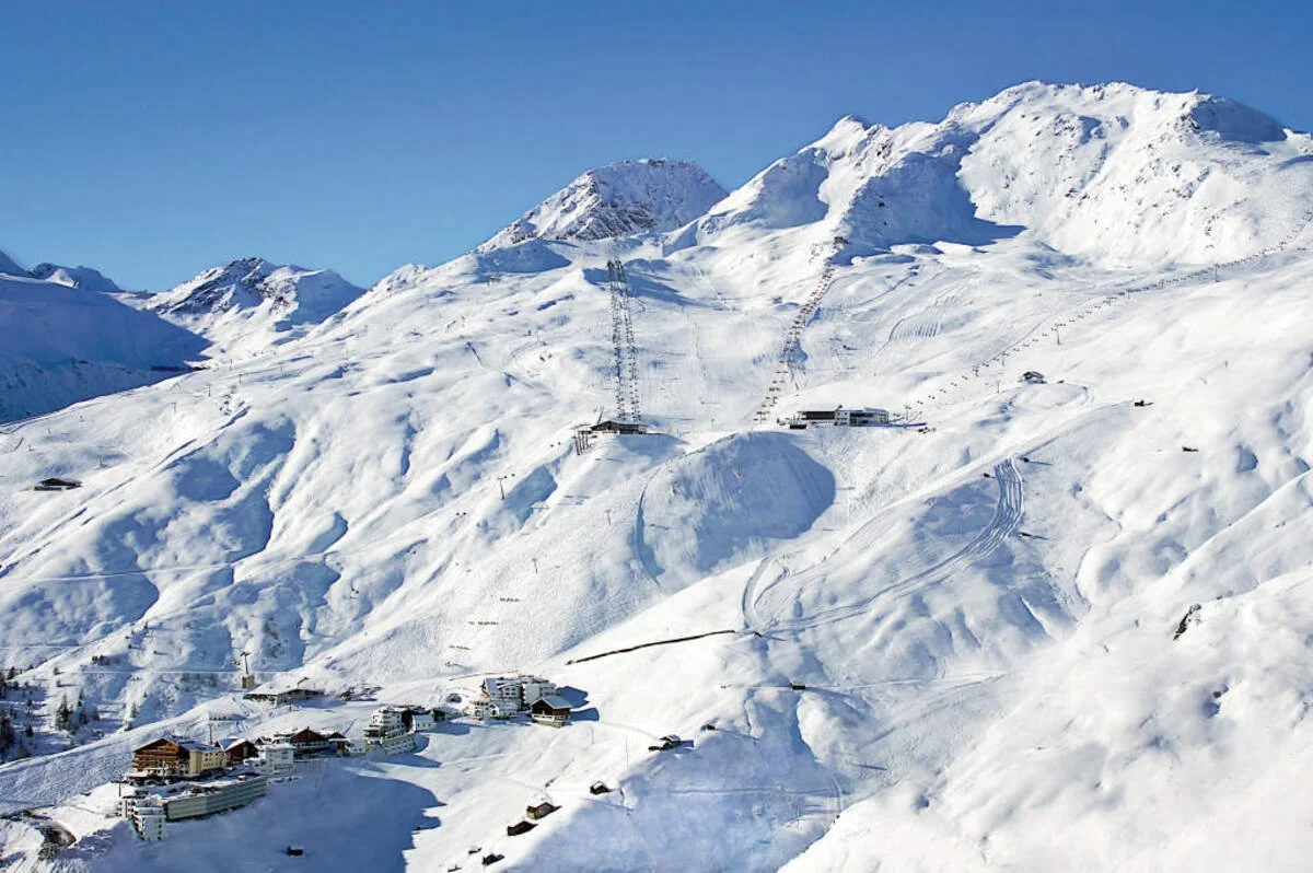 Snow-covered Solden mountains with ski slopes, ski lifts, and buildings at the base, under a clear blue sky.
