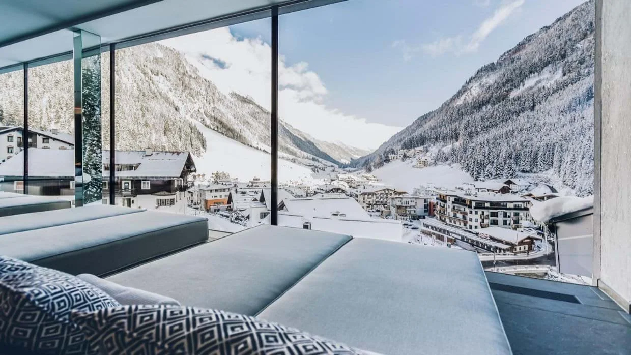 Indoor lounge with modern furniture and floor-to-ceiling windows offering a view of a snowy Ischgl mountain village and forested slopes.