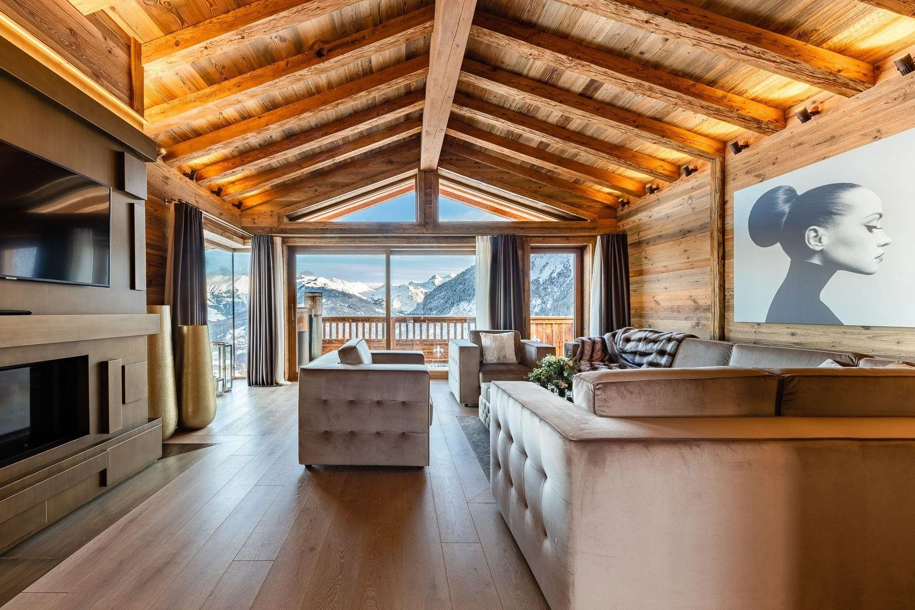 A cozy living room with wooden walls and ceiling, large windows showing snowy mountain views, and modern furniture including a sectional sofa, armchair, and a coffee table with flowers. There is also a black and white portrait of a woman on the wall.