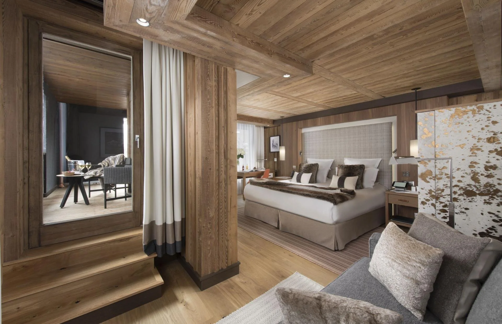 A cozy hotel room with wooden walls and ceiling, a large bed with multiple pillows, a bedside table with a phone, a lamp, and decorative items. There is a sitting area with a gray armchair and pillows, and a sliding door leading to a balcony with out