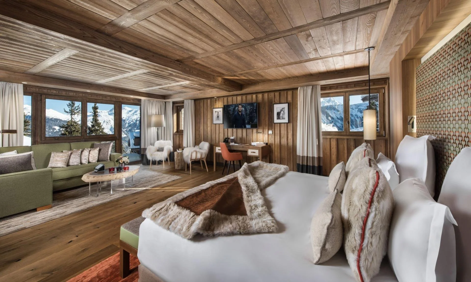 A cozy wooden cabin living room with large windows showing snowy mountain scenery; includes a bed with fur pillows, a green sofa with decorative pillows, a coffee table, a TV on the wall, and a workspace with a desk and chair.