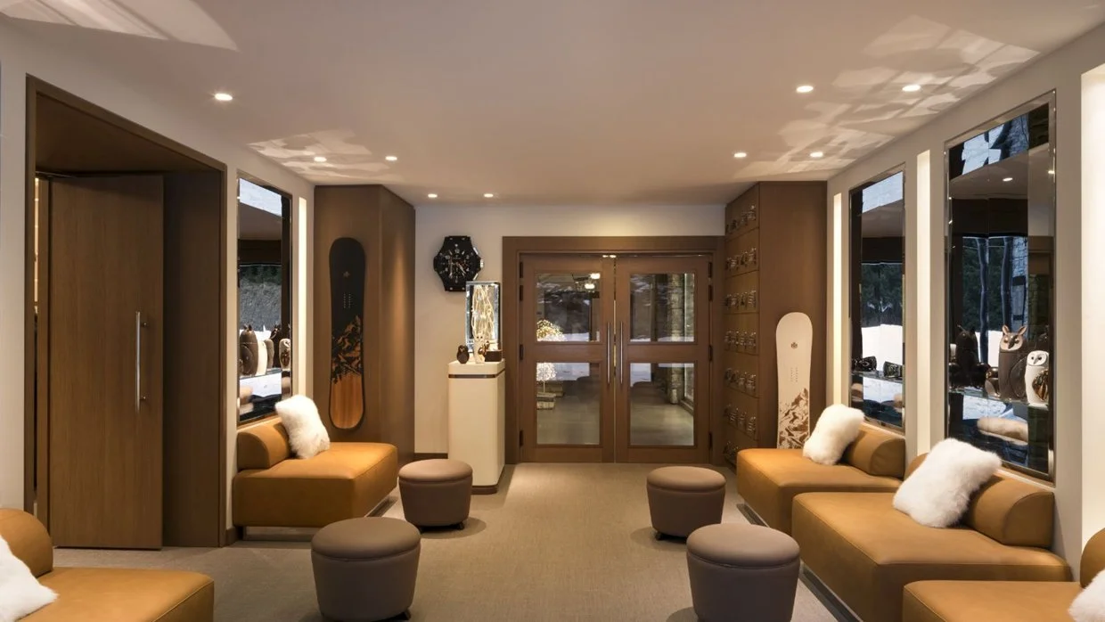 Interior of a modern lounge area with brown sofas, white pillows, and round ottomans. There are wooden sliding doors at the back, large windows with display shelves showing decorative items, and decorative skateboards on the walls.