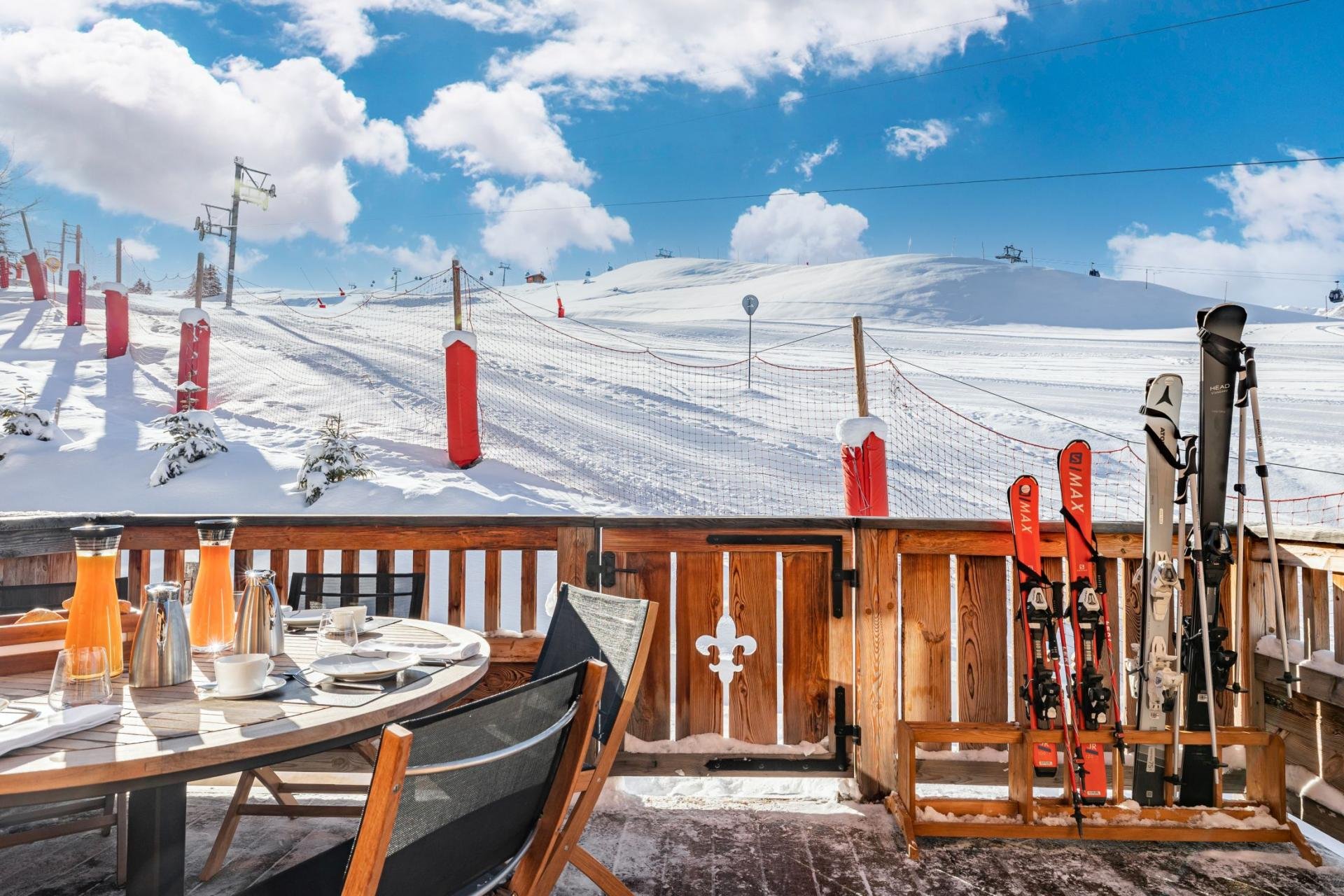 A snow-covered ski resort viewed from a wooden deck with a table set for breakfast and ski equipment, including skis and poles, arranged near the deck railing, under a bright blue sky with scattered clouds.