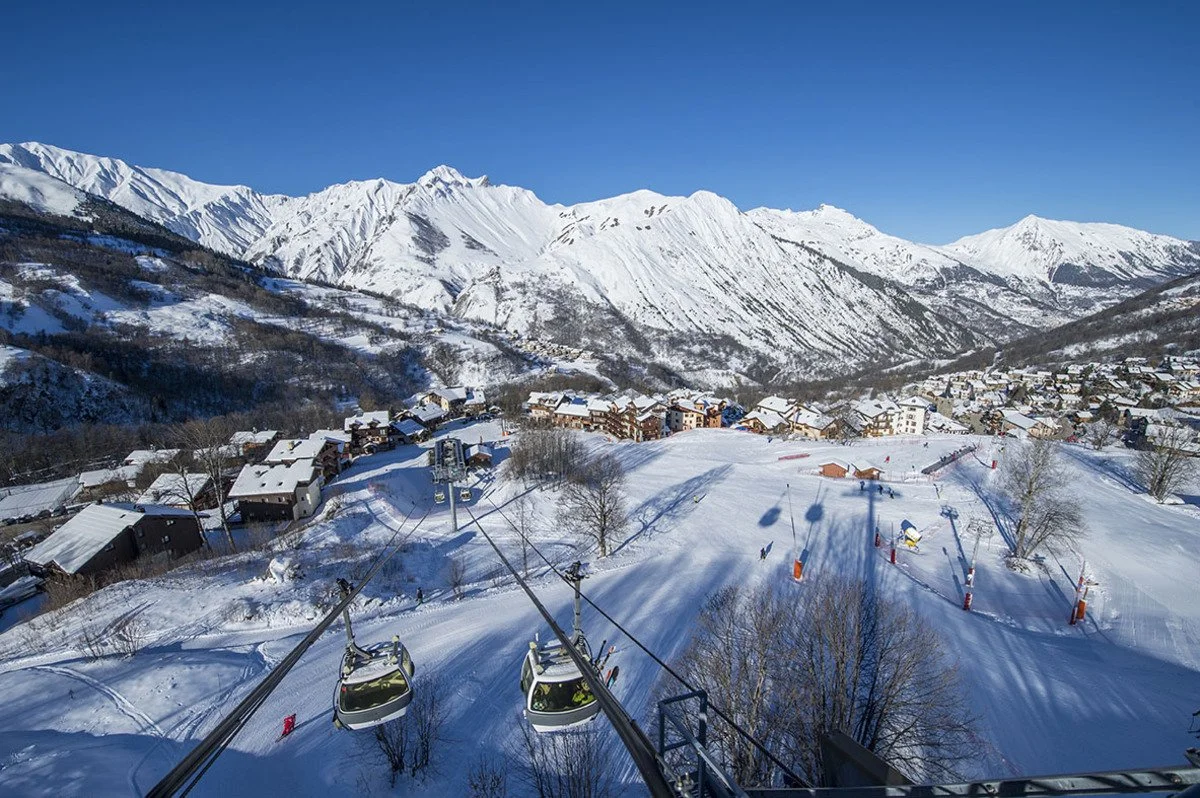 View of a snowy St Martin de Belleville ski resort village with chairlifts, snow-covered buildings, and mountains in the background on a clear blue sky day.