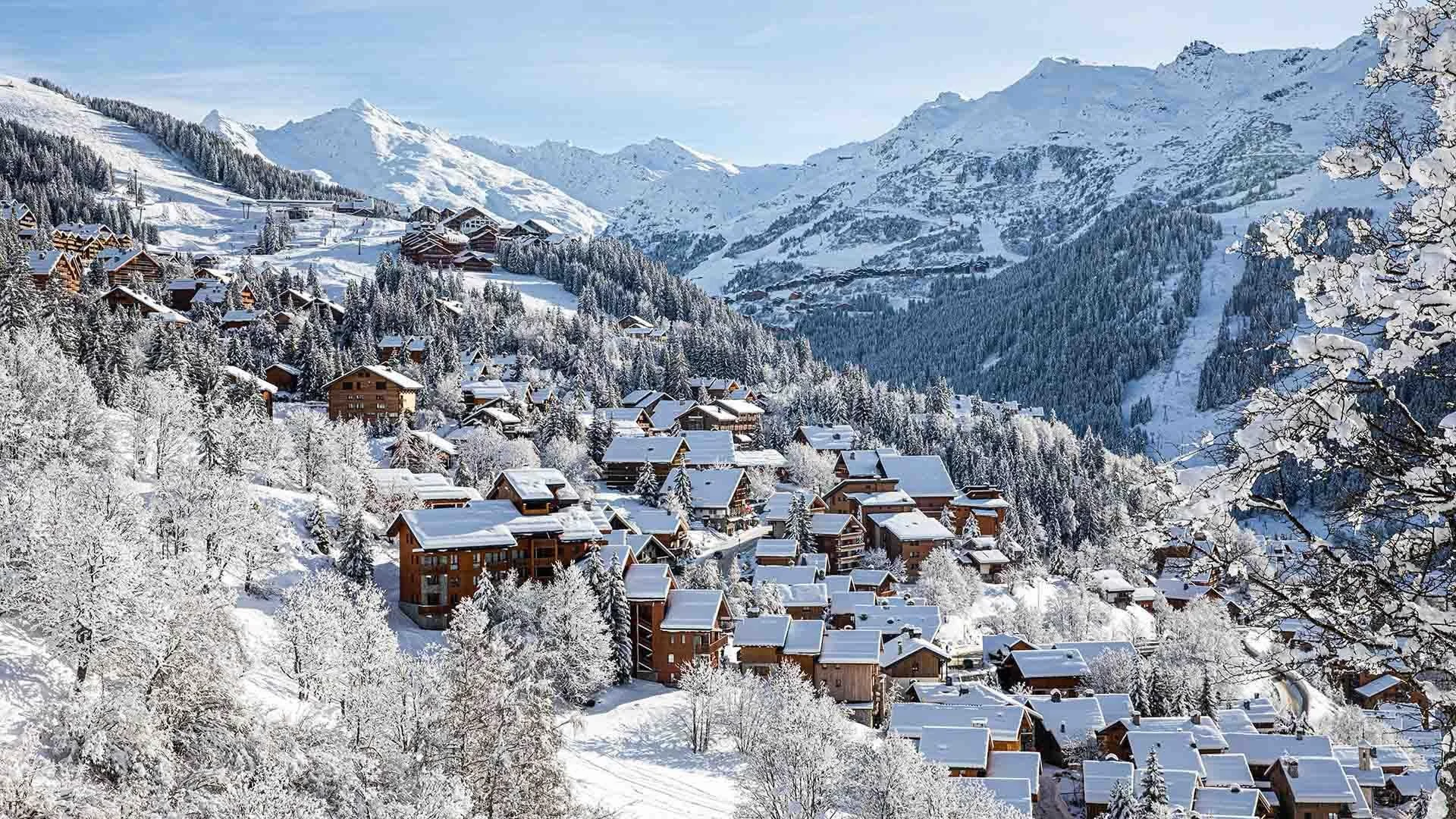Snow-covered Meribel mountain village with wooden houses, surrounded by snow-laden trees and high snow-capped mountain peaks in the background.