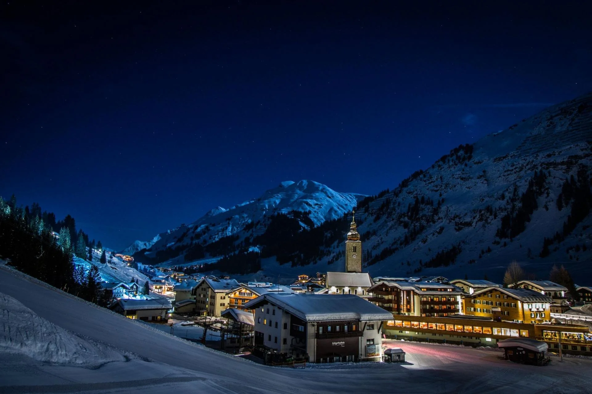 Night view of a snowy Lech mountain village with illuminated buildings and a church steeple, surrounded by snow-covered mountains under a starry sky.