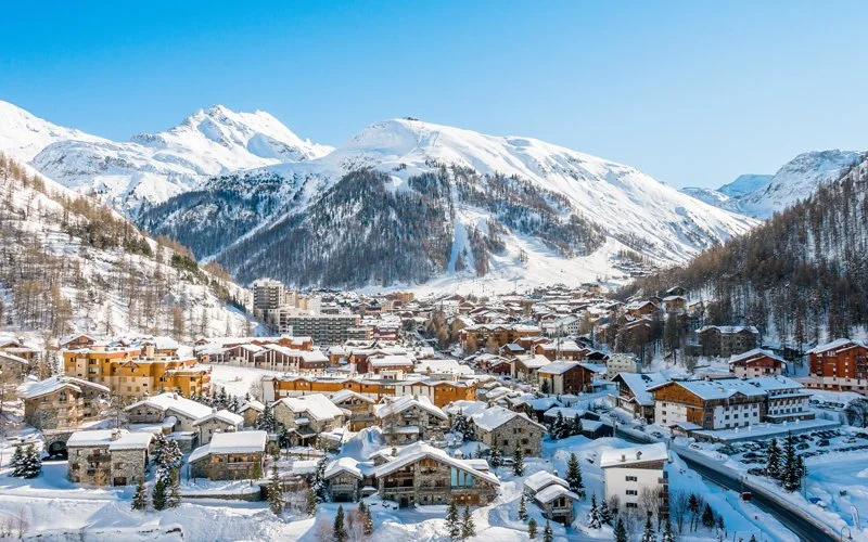 Snow-covered Val d'Isere town nestled in a valley surrounded by tall, snow-capped mountains under a clear blue sky.