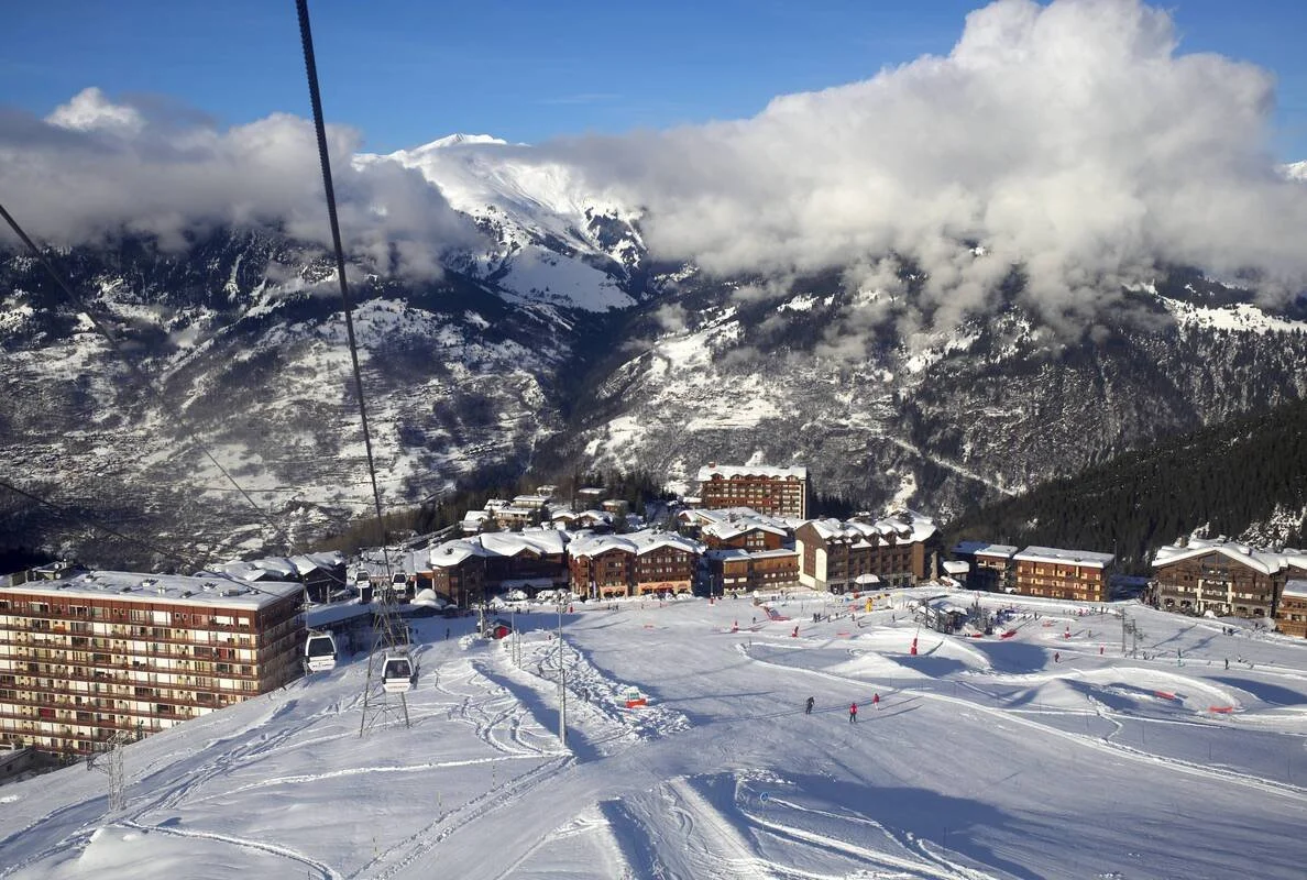 A snowy Courchevel 1650 mountain resort with ski slopes, buildings, and ski lift cables amid mountains and cloudy sky.