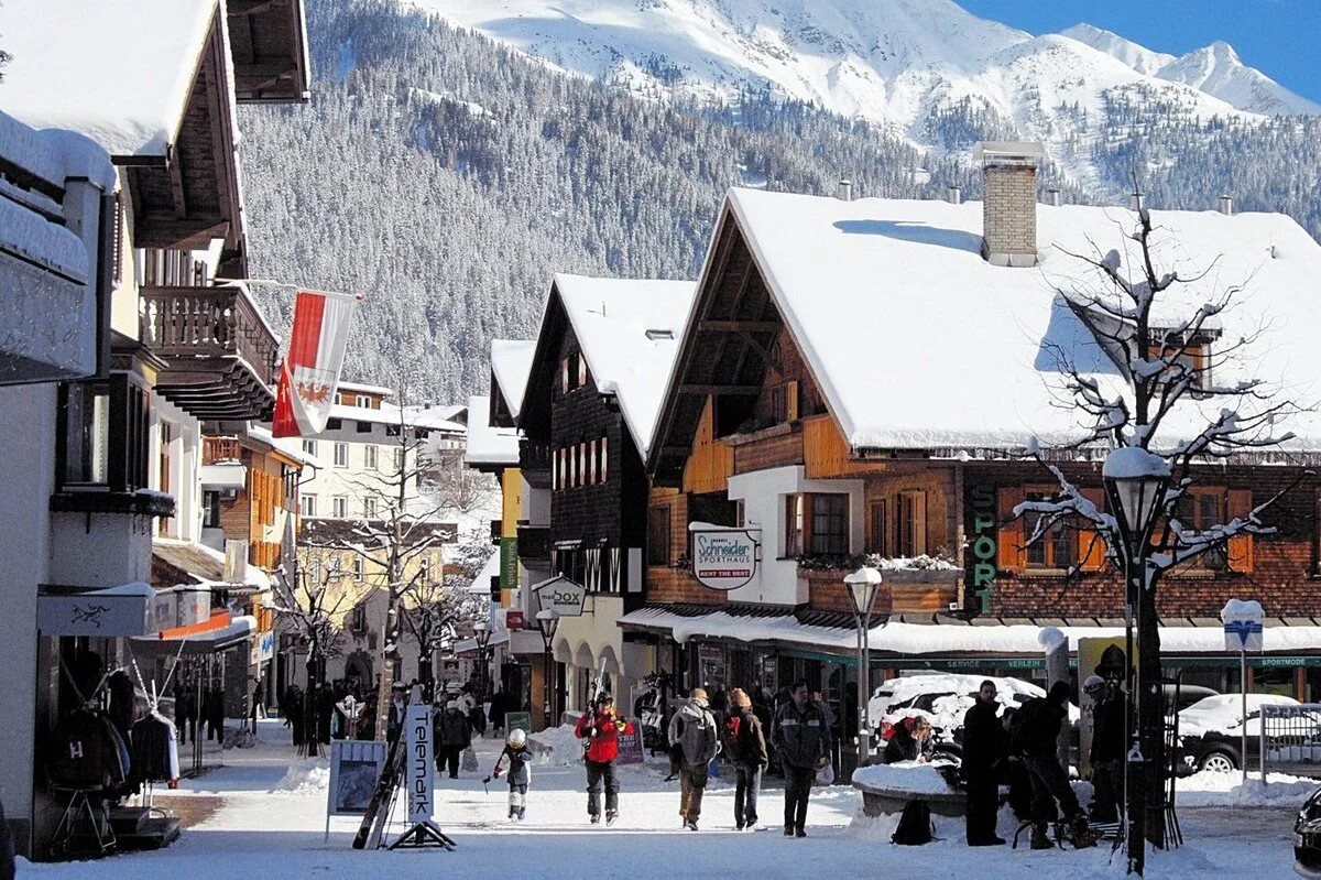 A snowy mountain village scene with pedestrians walking on a snow-covered street lined with shops and buildings. Snow is on rooftops and trees, with mountains in the background.
