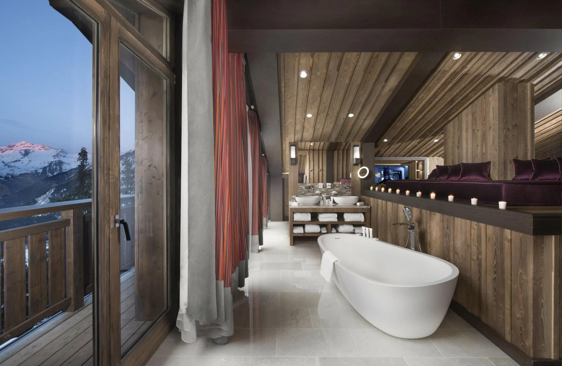 A modern bathroom with wooden walls and ceiling, a freestanding bathtub, a sink with a large mirror, candles, and a balcony with a mountain view.