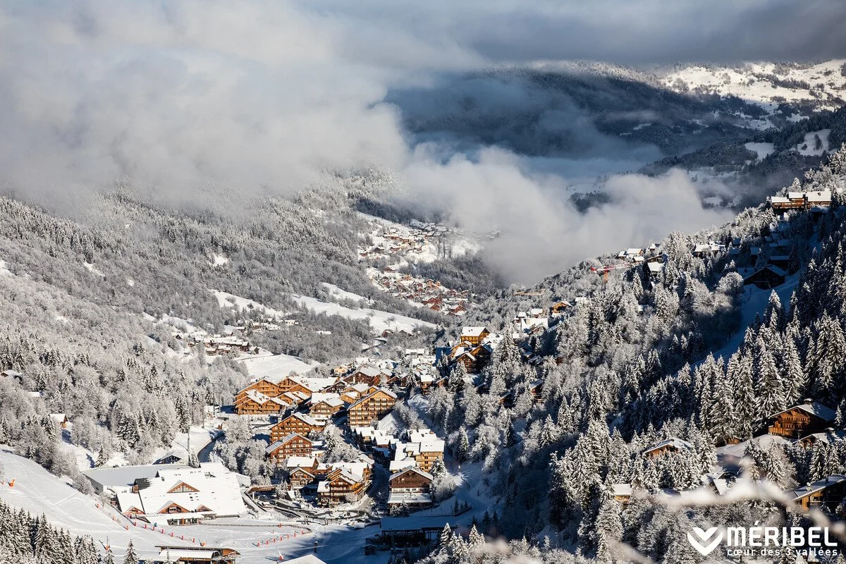 Snow-covered Meribel village with chalets and houses, dense pine trees blanketed in snow, fog and clouds in the background, sunny sky