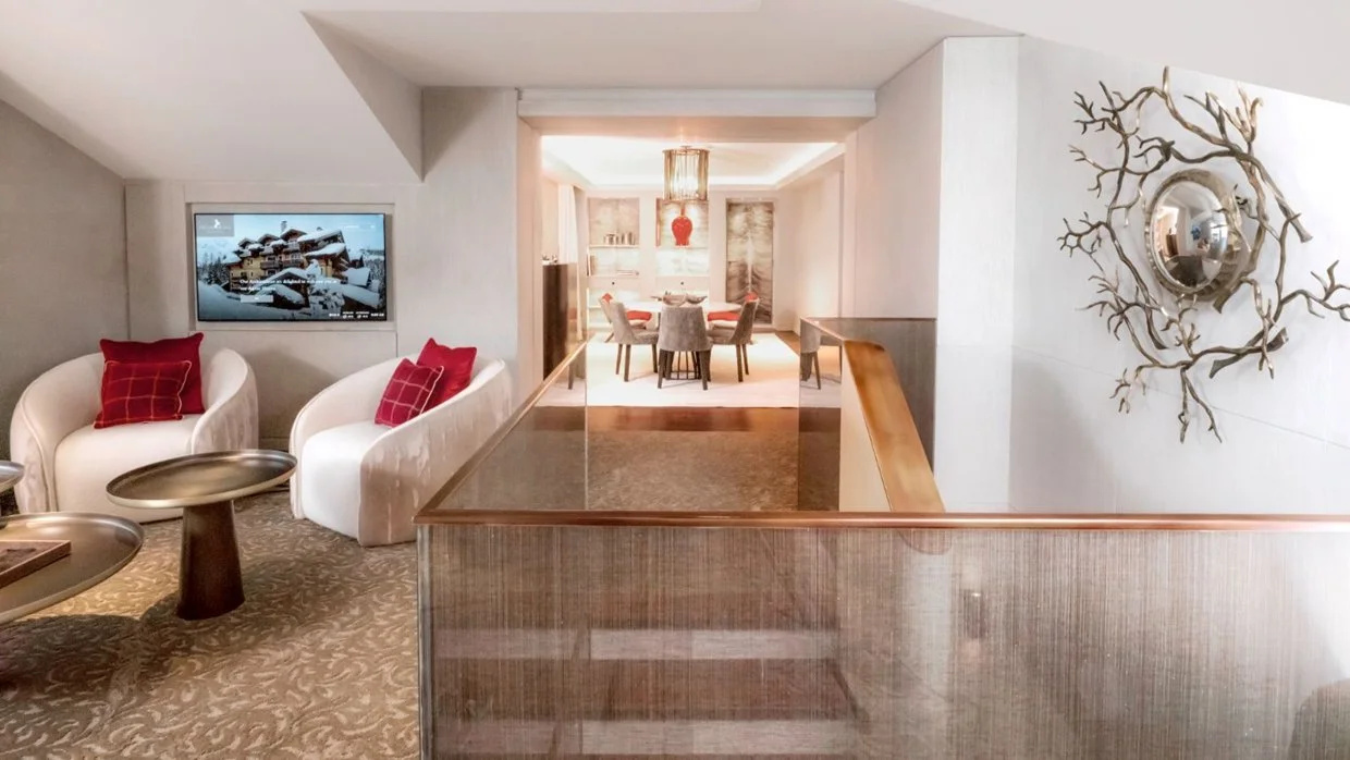 Living room with white armchairs, red pillows, wall-mounted TV showing a snowy landscape, decorative wall mirror with tree branch design, and a dining area in the background.
