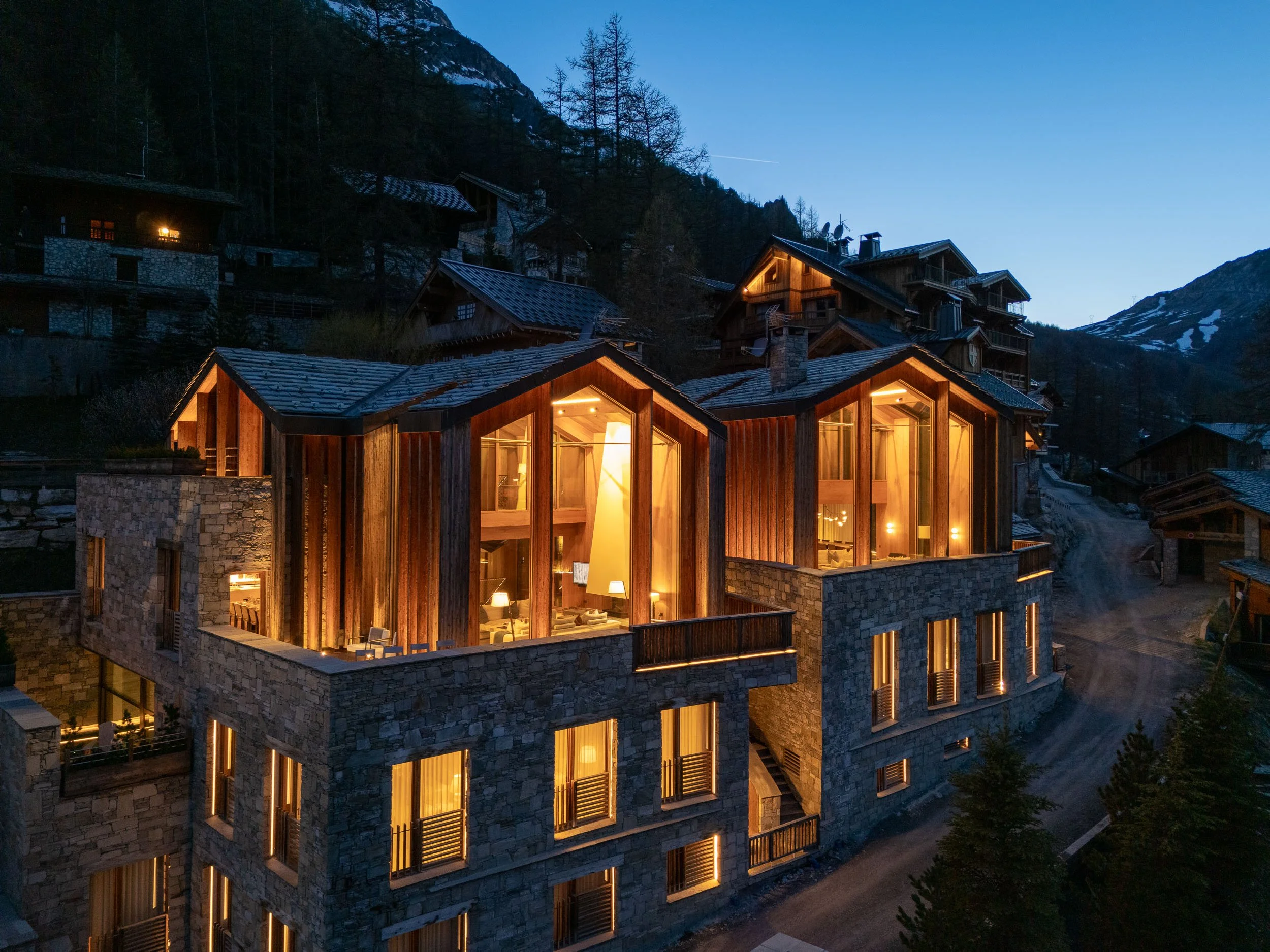 An illuminated modern multi-story mountain chalet at dusk with large glass windows and wooden accents, surrounded by pines and mountain terrain.