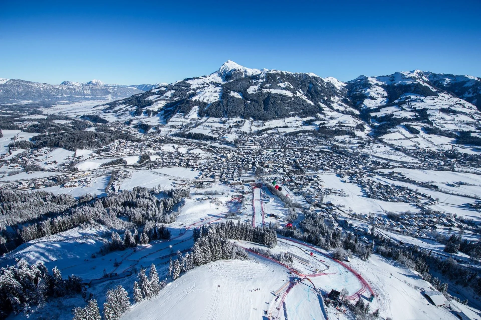 Aerial view of a snowy mountain landscape featuring Kitzbuhel ski resort, with ski slopes, chairlifts, and a nearby town against a backdrop of snow-covered mountains and a clear blue sky.