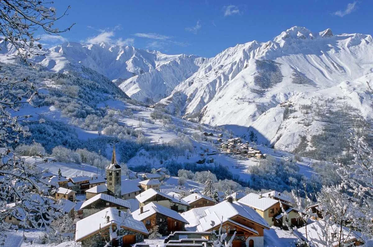 A snow-covered St Martin de Belleville village in a mountainous winter landscape with snow-capped peaks, ski slopes, and a clear blue sky.