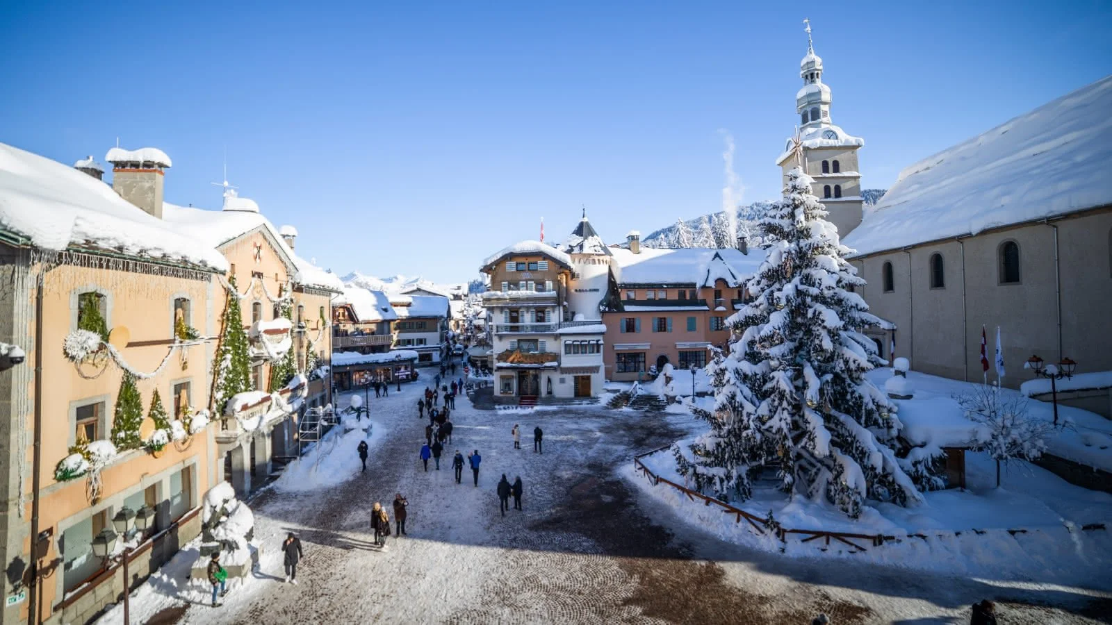 Snow-covered Megeve town square with people walking, decorated buildings, and a large snow-laden Christmas tree, with a church tower in the background under a clear blue sky.
