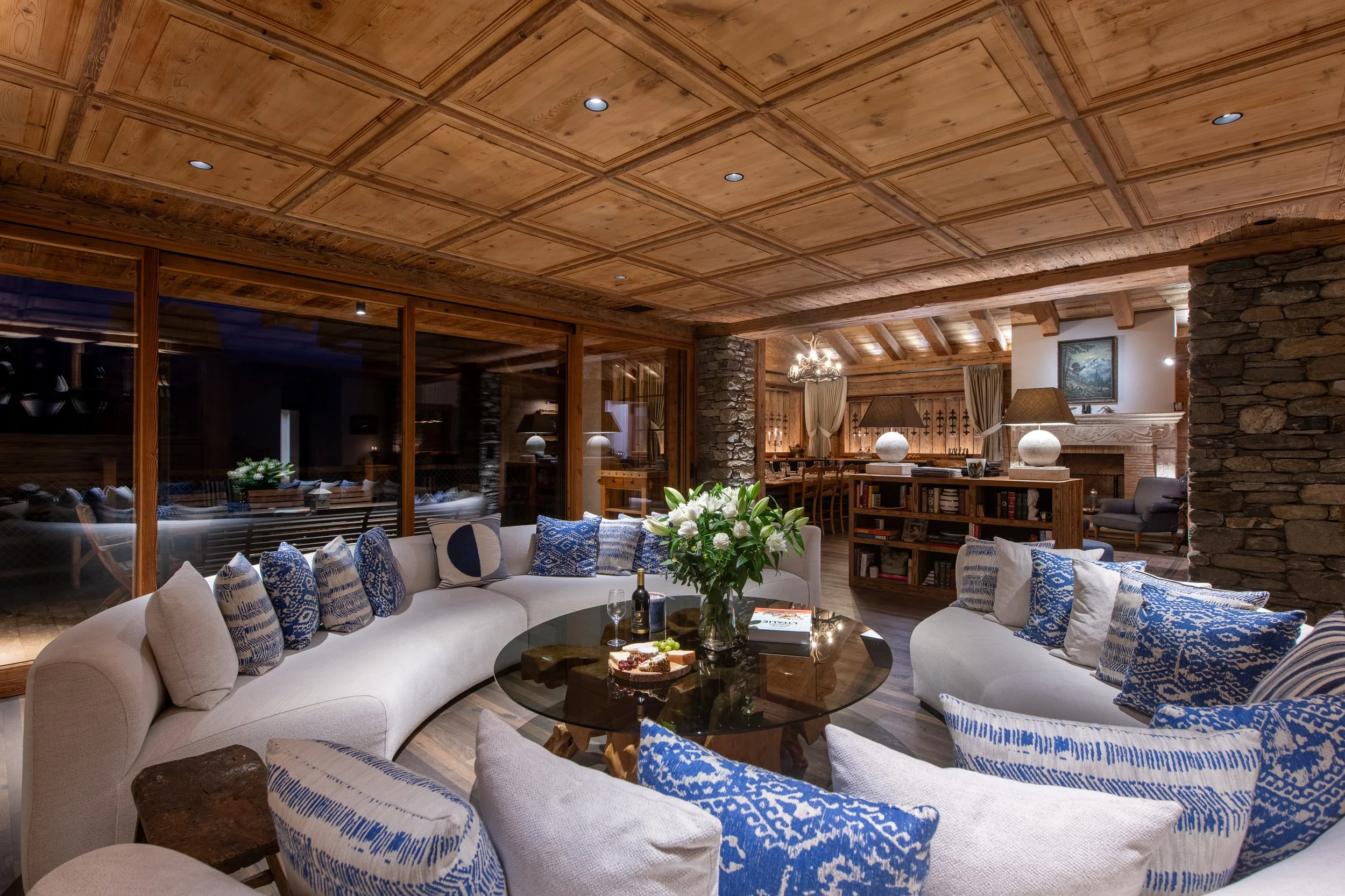 Living room with wooden paneled ceiling, stone walls, and cozy seating with blue and white patterned cushions. There is a glass coffee table with snacks and drinks, and a floral centerpiece. The space features a stone fireplace, bookshelves, and warm lighting.