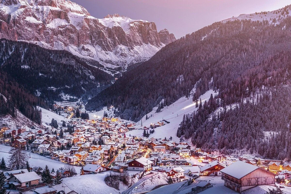 Snow-covered mountain village in a valley surrounded by tall, snow-capped mountains at dusk, with the village lights beginning to turn on.