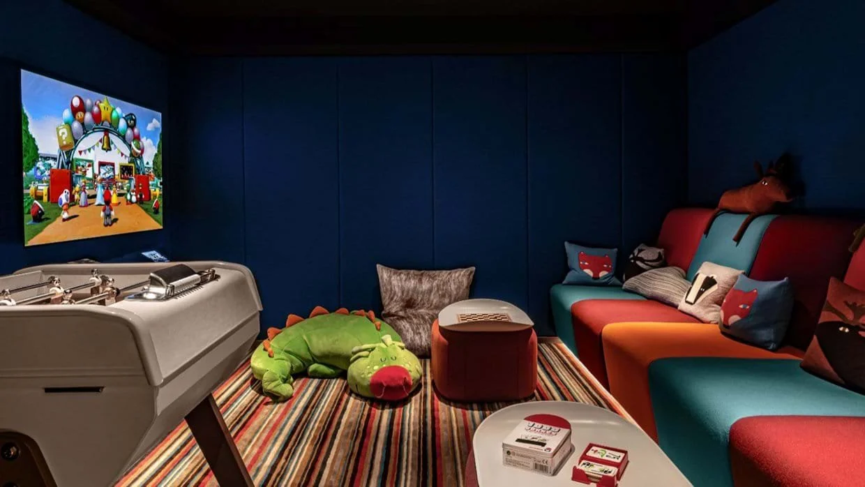 A colorful game room with a TV displaying a Mario-themed game, a foosball table, a plush green dinosaur toy, a cozy gray pillow, a red and green dinosaur pillow, a small white table with a card game box, and a variety of vibrant sofas with playful pi
