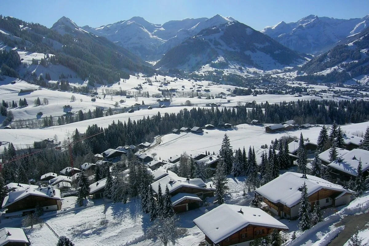 Snow-covered Gstaad village in a valley surrounded by mountains with ski slopes and houses with snow-covered roofs