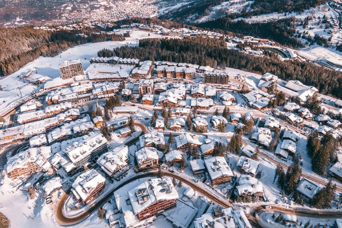 Aerial view of a snow-covered Courchevel 1850 with houses, roads, and forested hills in the background.