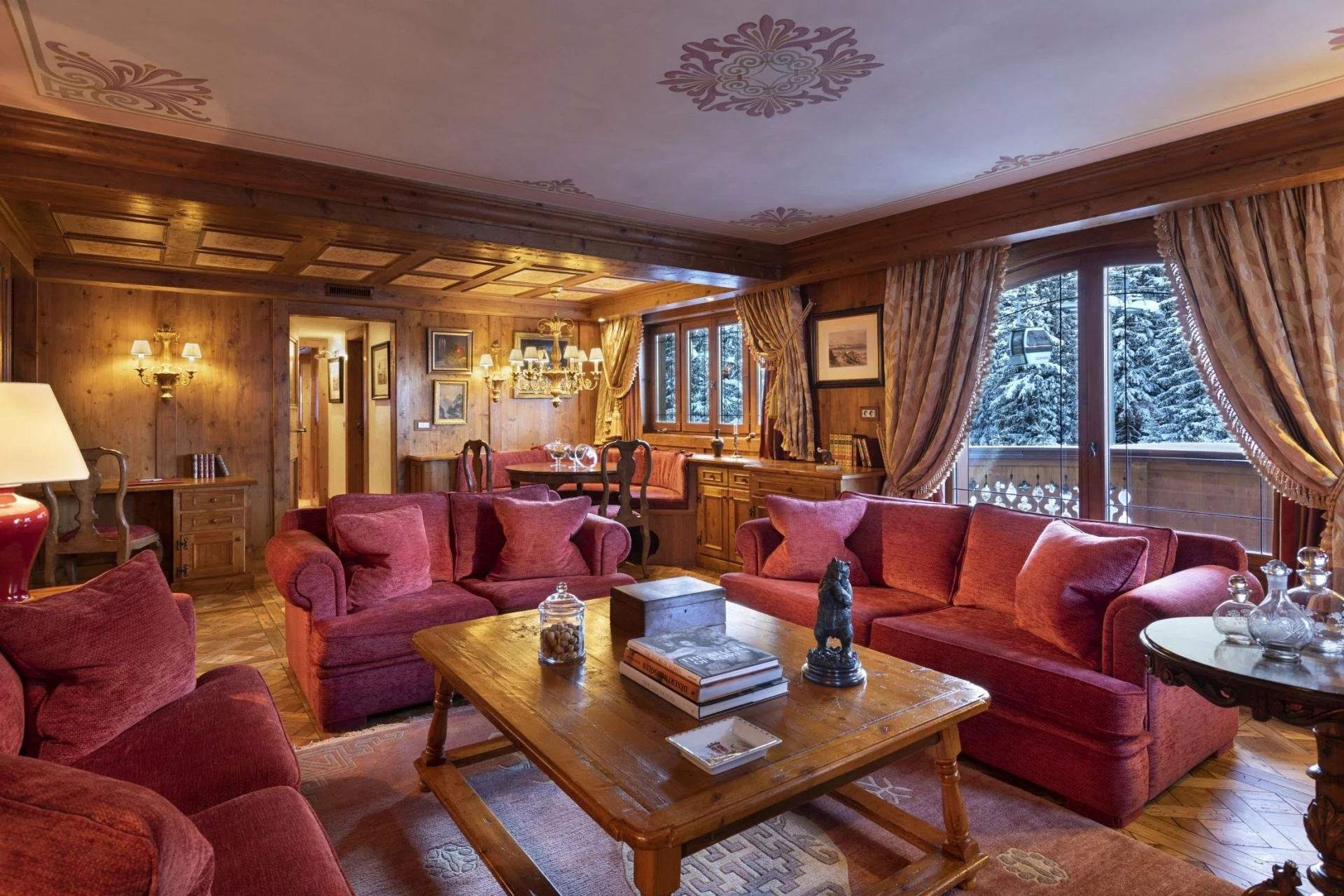 A cozy living room with wooden walls and ceiling, large windows showing a snowy outdoor landscape, red sofas, wooden furniture, and elegant lighting fixtures.