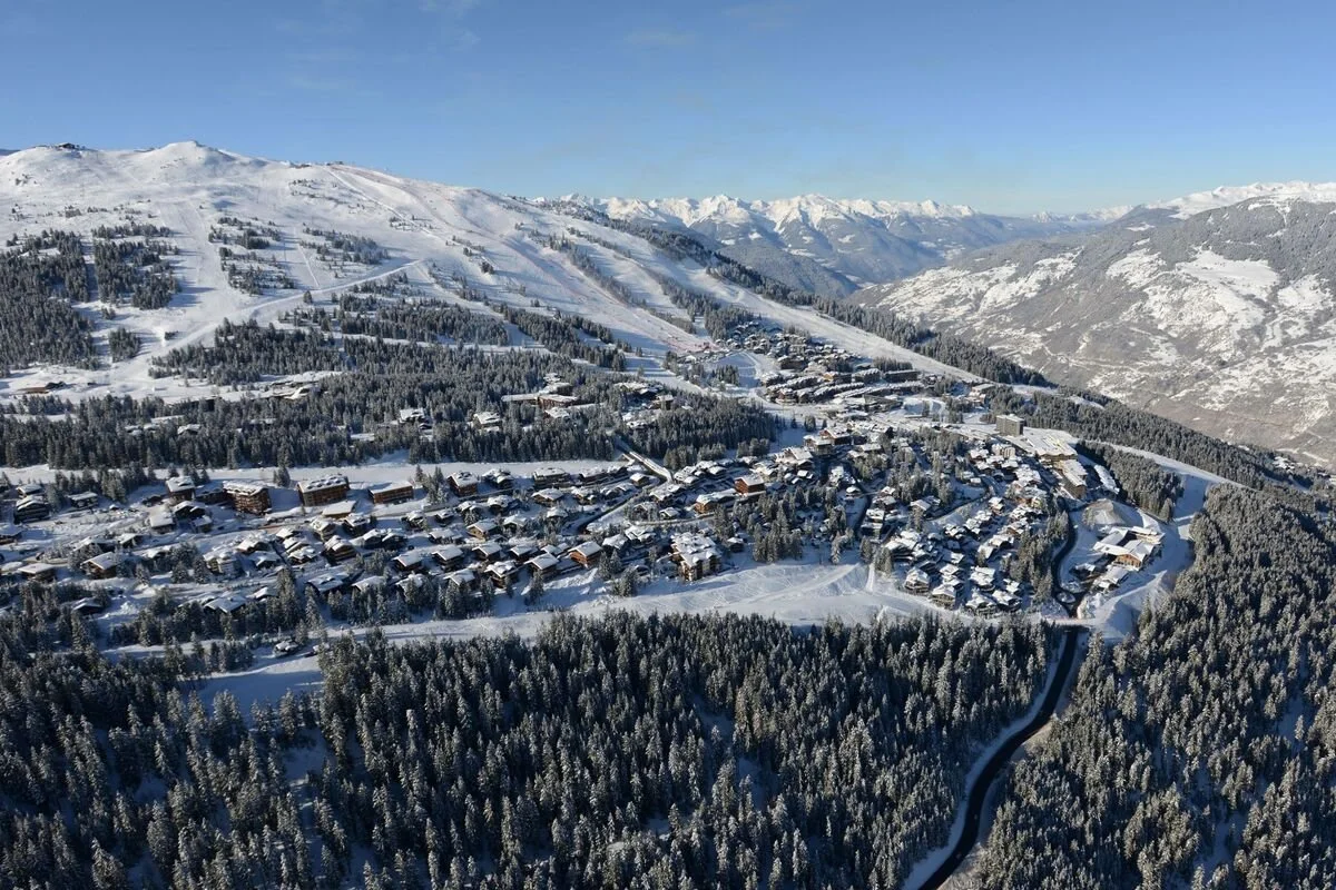 A snow-covered Courchevel 1850 village with many houses, surrounded by dense snow-covered forest and mountain slopes in the background.