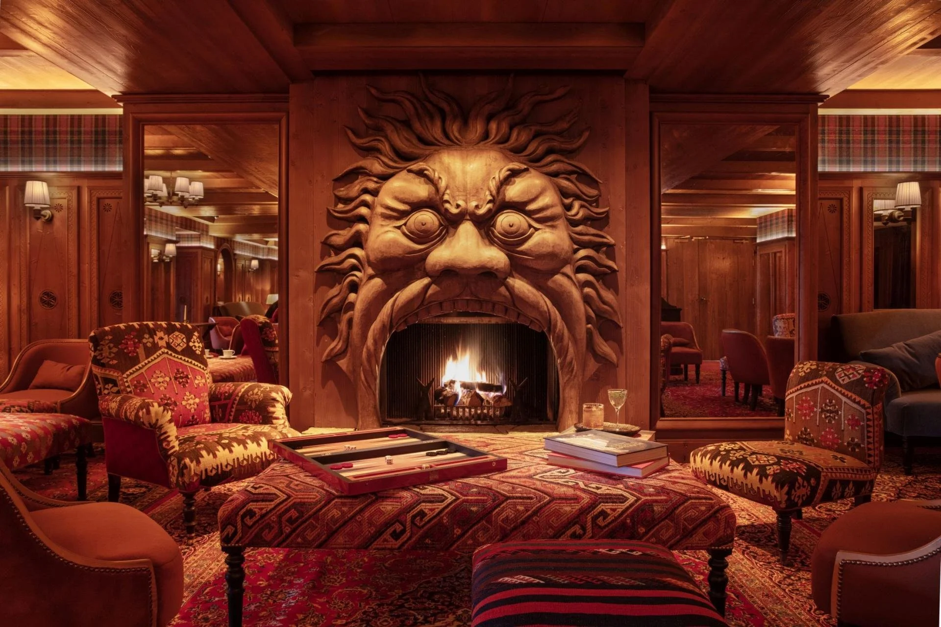 A cozy lounge with a carved lion's head fireplace and a fire burning. The room has wooden paneling, patterned armchairs, and selvedge plaid accents.