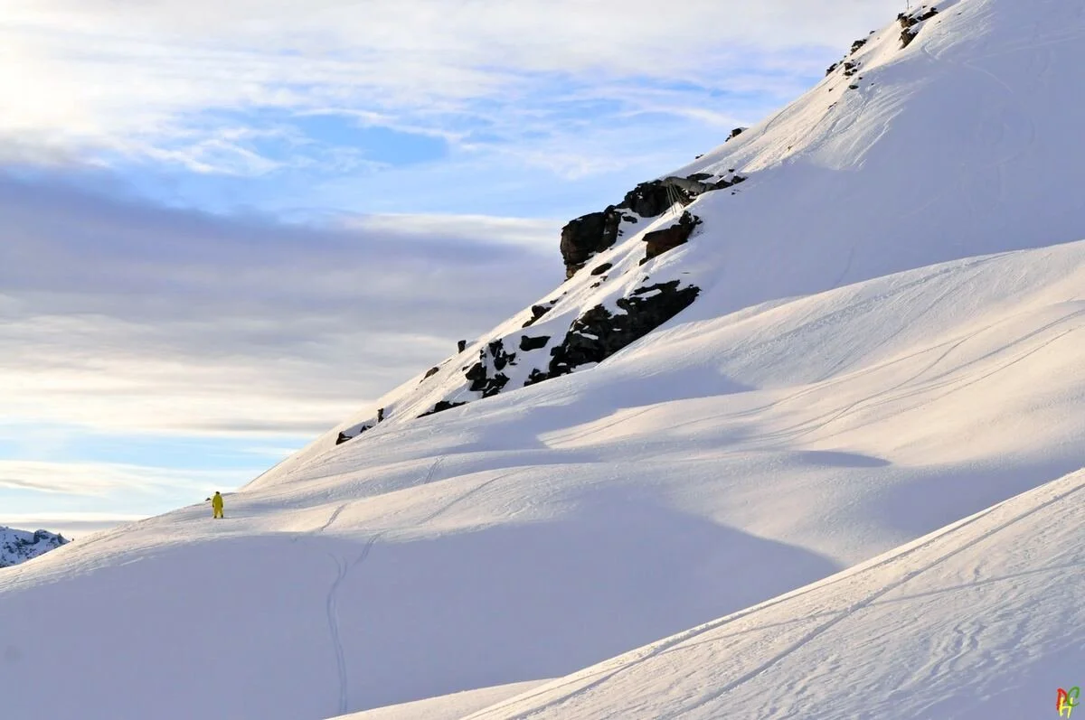 A snowy mountain slope in Val Thorens with a lone skier dressed in yellow gear, near rocky outcroppings, under a partly cloudy sky.