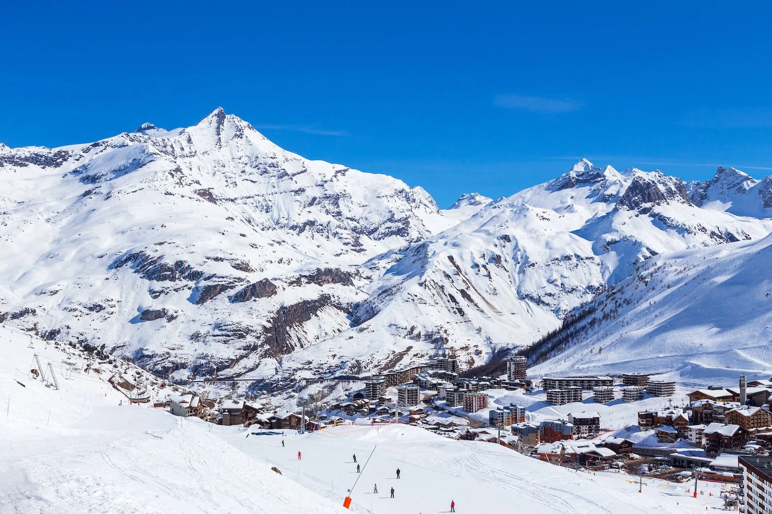 Snow-covered Tignes resort town in the mountains with skiers on the slopes and buildings at the base, under a bright blue sky.