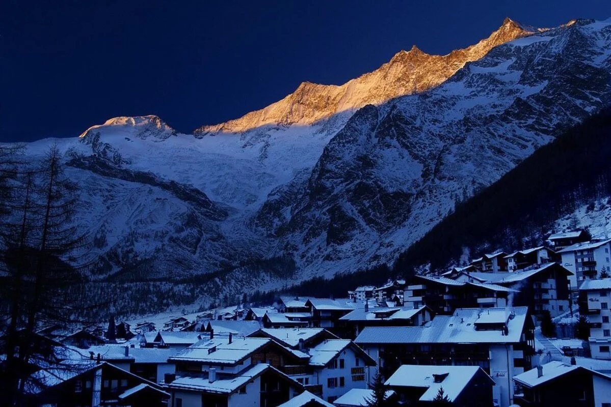 Snow-covered Saas-Fee alpine village with dark rooftops below tall, snow-capped mountains during sunrise or sunset.