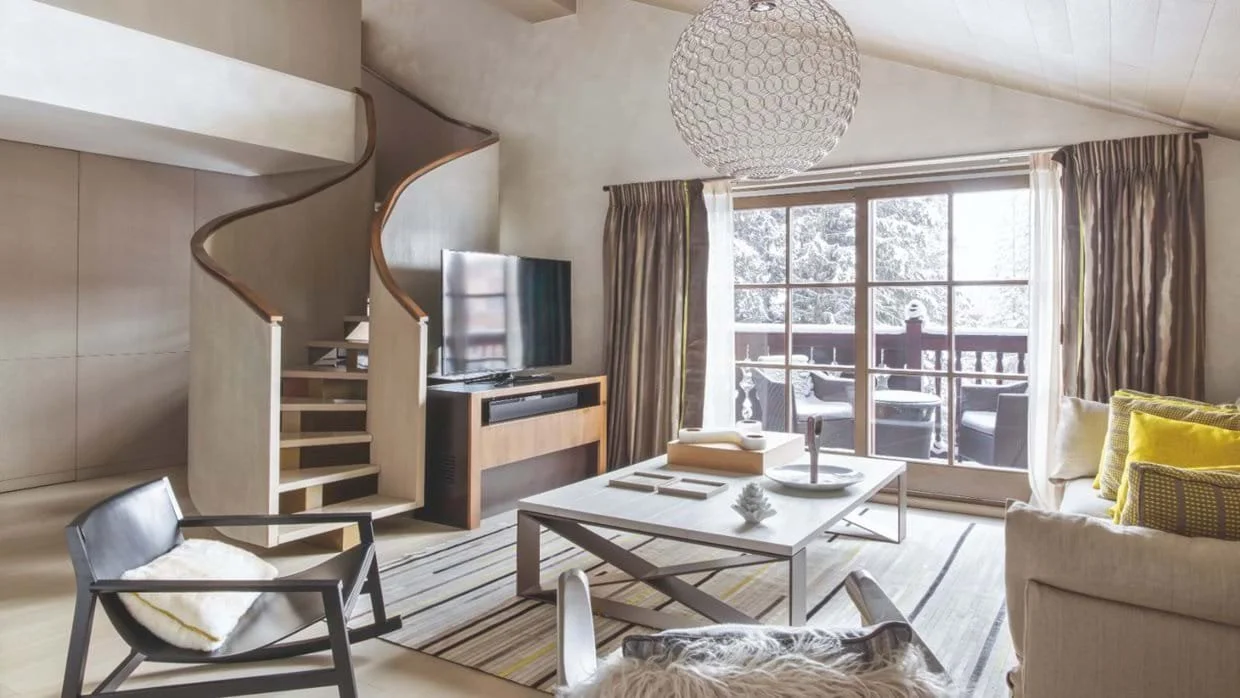 Living room with beige walls, large window leading to a balcony with snow-covered trees, a spiral staircase, a fireplace, a flat-screen TV, a coffee table with decor, a sofa with yellow and patterned cushions, a black chair with a blanket, and a roun
