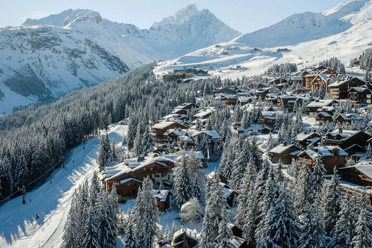 Snow-covered Courchevel 1850 village with wooden chalets and tall pine trees, surrounded by snow-capped peaks.