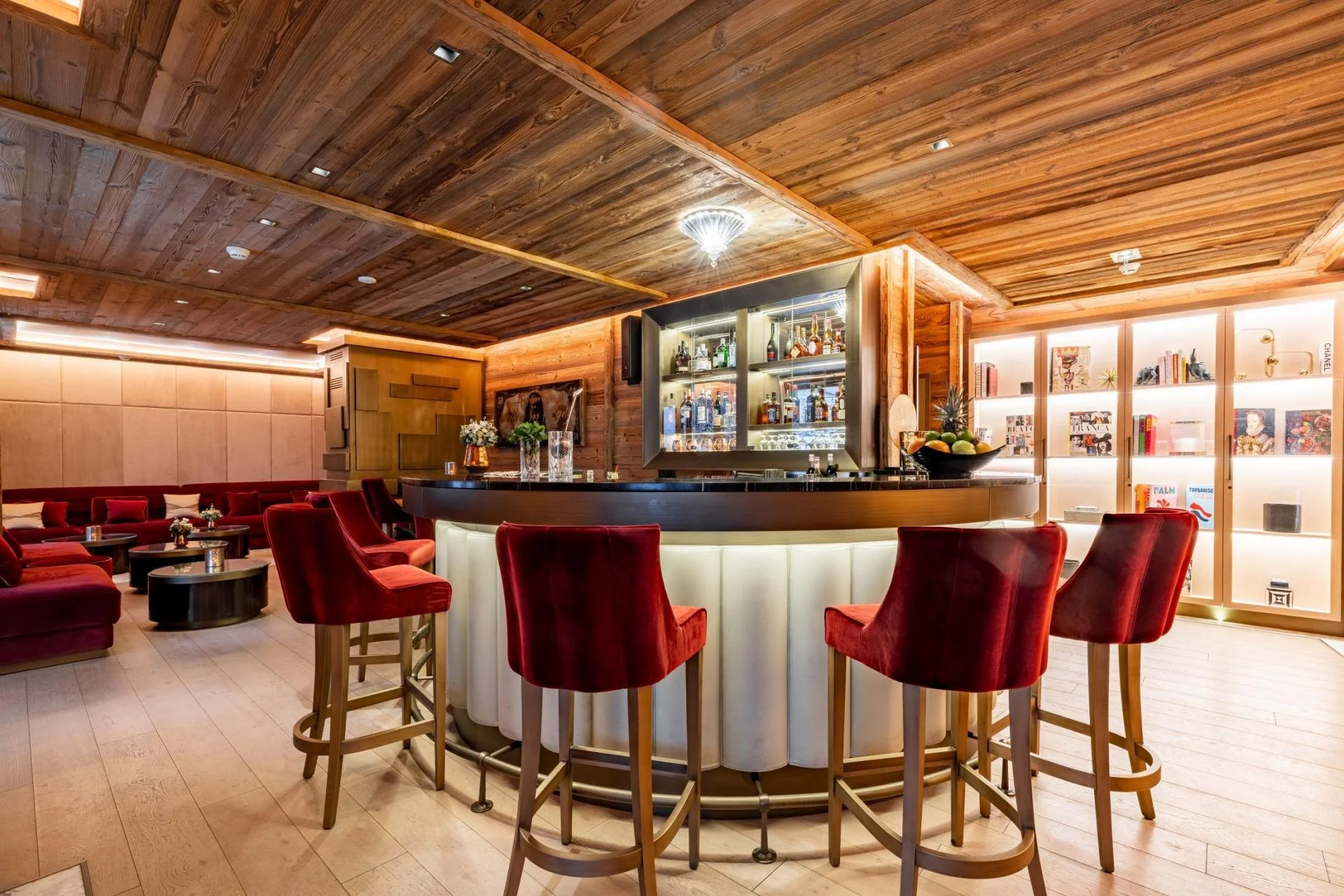 Interior of a bar area with a curved bar counter, red chairs, a wooden ceiling, and illuminated shelving with liquor bottles.