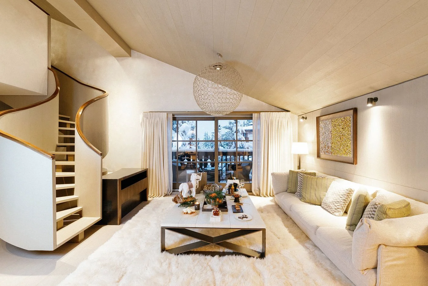 A cozy living room with a staircase, a large cream sofa with throw pillows, a coffee table with snacks, and sliding glass doors leading to a snowy outdoor deck.