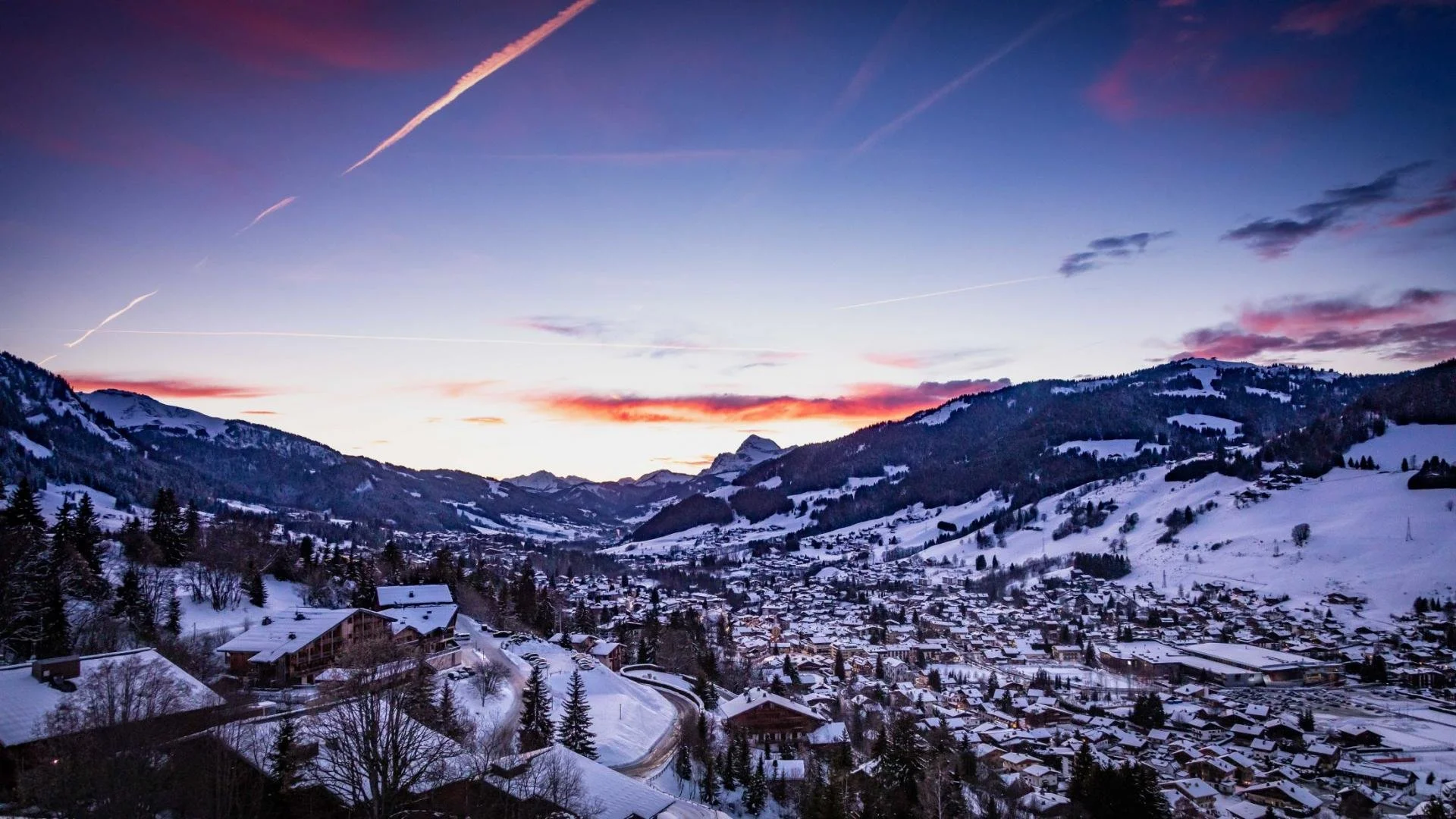Snow-covered Megeve village in a valley surrounded by mountains during sunset, with pink and purple sky and visible contrails.