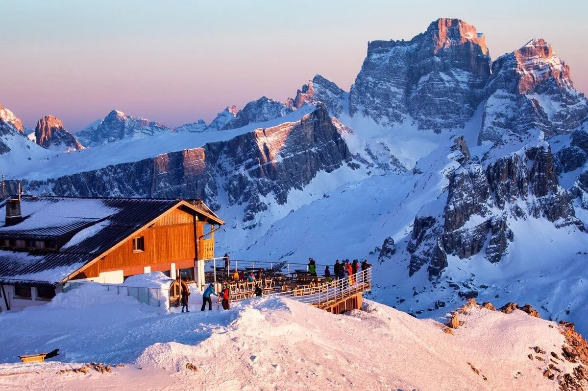 Snow-covered mountain landscape with a wooden chalet and people on a balcony, mountain peaks in the background during sunset.