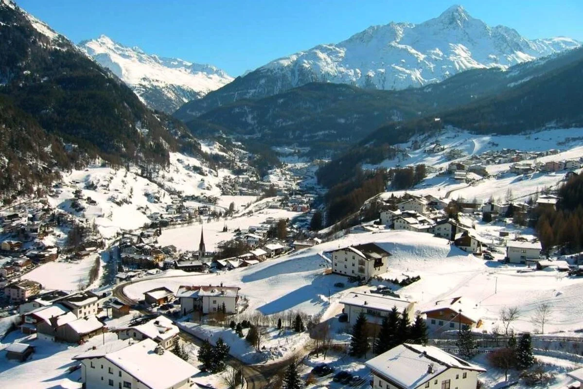 Snow-covered Solden mountain village with houses, trees, and a church steeple, surrounded by snow-capped mountains under a clear blue sky.