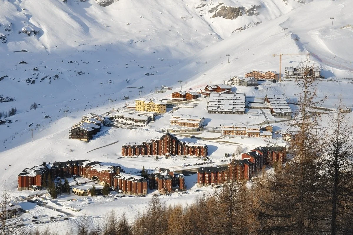 Snow-covered mountains with a clustered ski resort town consisting of several buildings and houses, ski lifts and construction cranes visible.