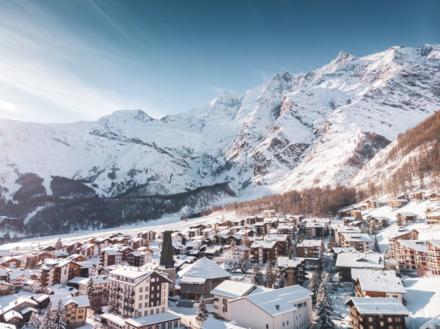 Snow-covered Saas-Fee alpine village with tall mountains in the background under a clear blue sky.