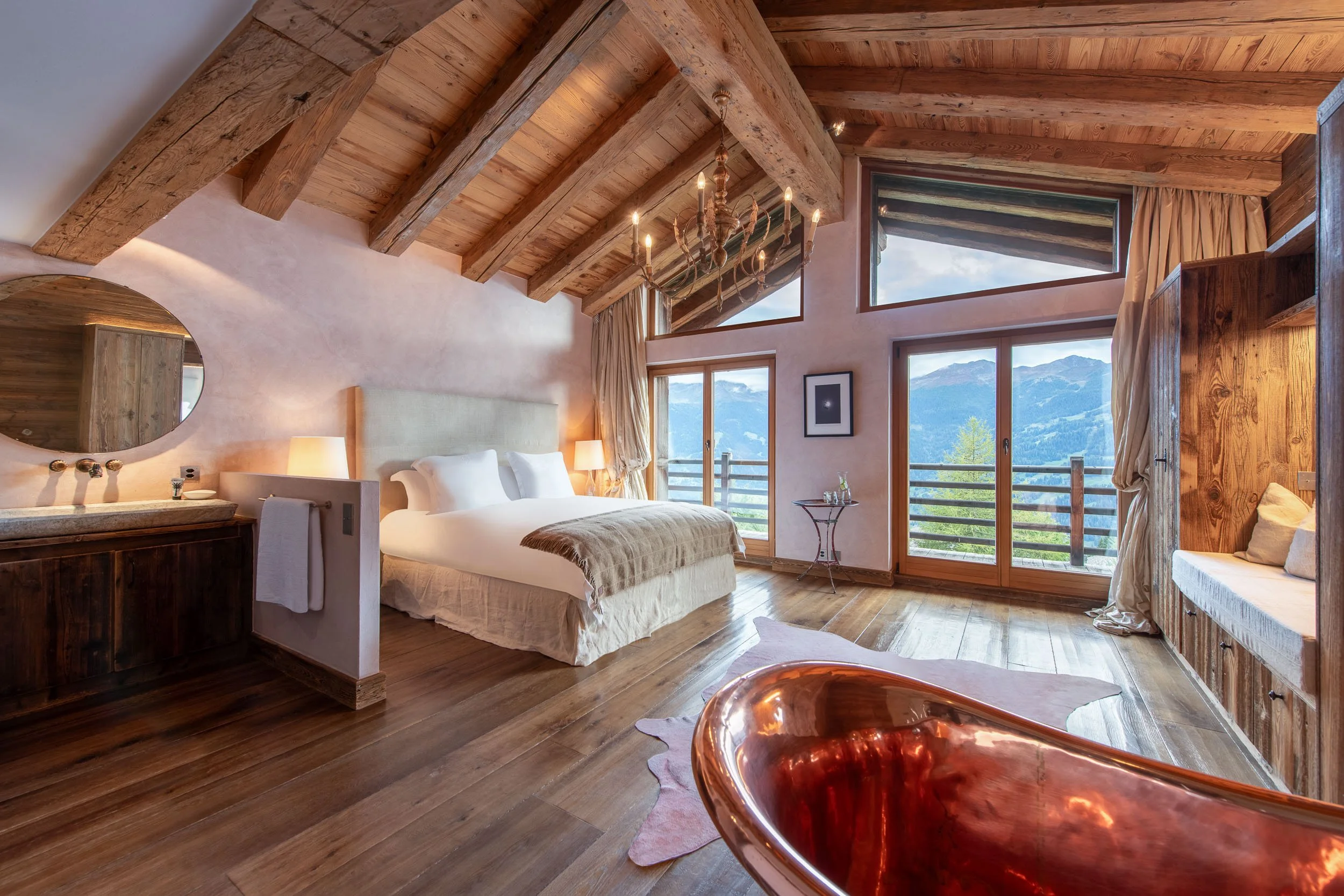 A cozy bedroom with a wooden vaulted ceiling, large windows showing mountain views, a king-sized bed with white bedding, a chandelier, and rustic furniture including a copper bathtub and wooden wardrobe.