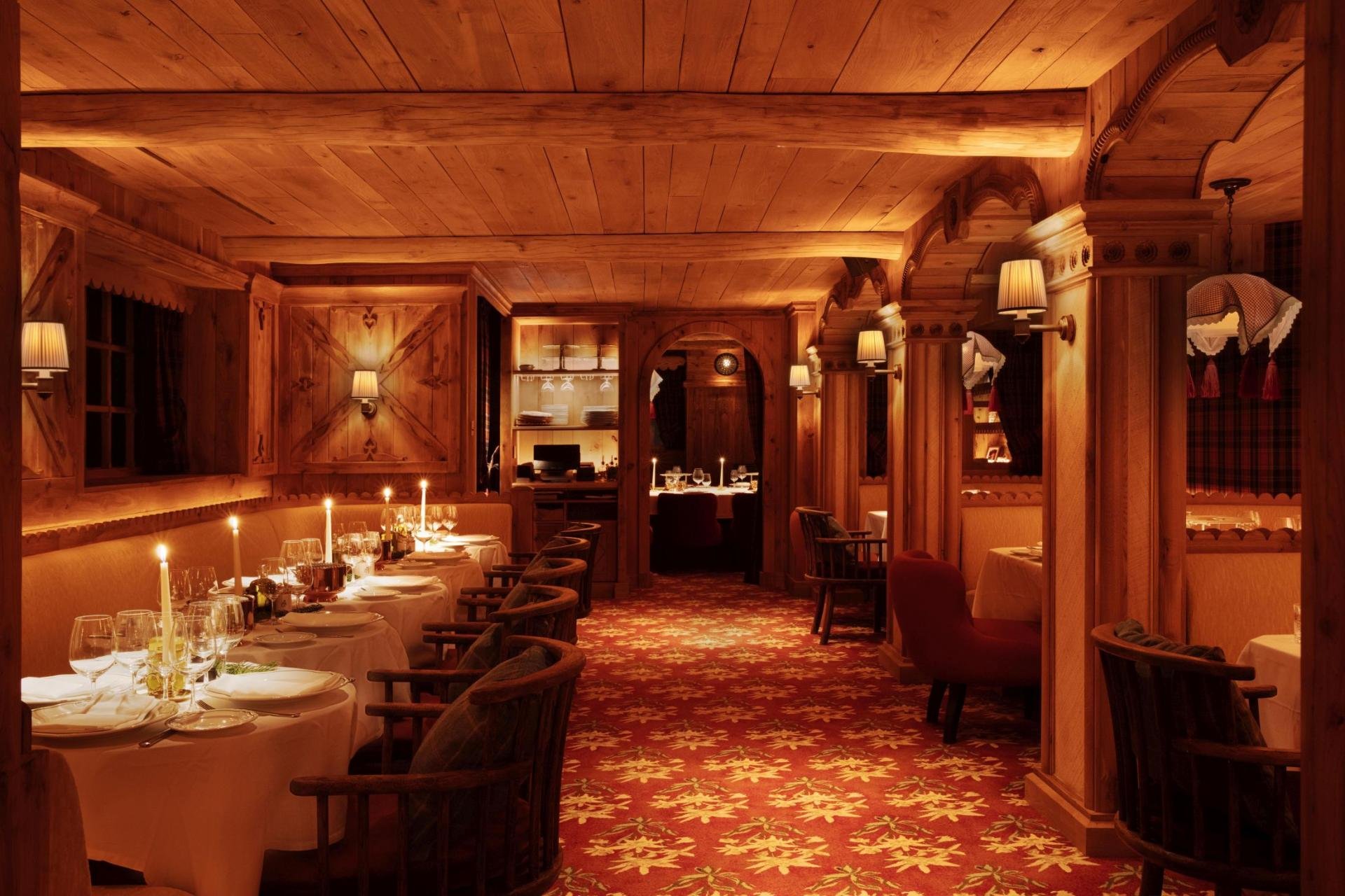 A cozy, warmly lit dining room with wooden walls and ceiling, round tables with white tablecloths, wine glasses, cutlery, lit candles, wooden chairs, and a patterned red carpet.