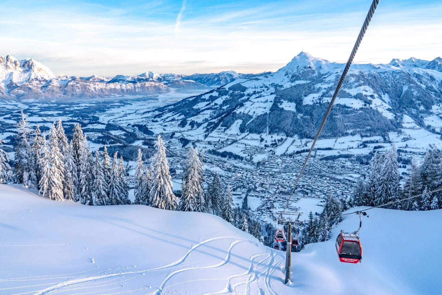 Snow-covered Kitzbuhel mountain landscape with a ski lift and a village below, surrounded by mountains and pine trees.