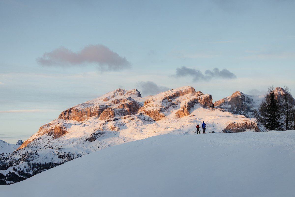 Two people hiking in snowy mountains during sunset with a partly cloudy sky.