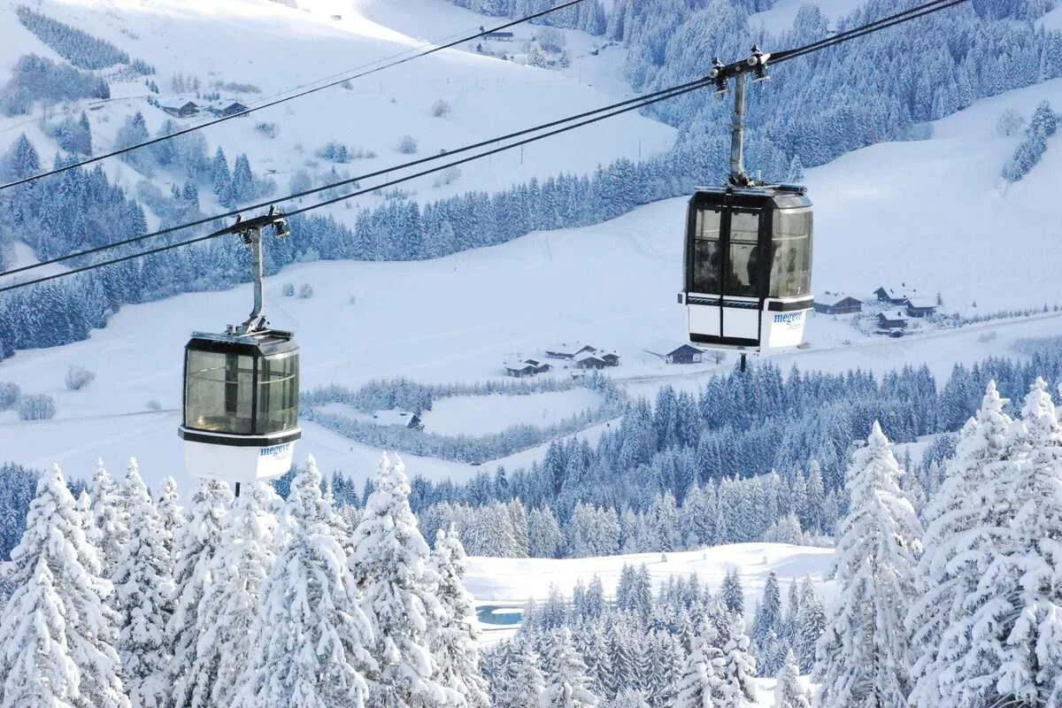 Two ski lifts in Megeve with enclosed cabins traveling over snow-covered trees and a mountainous landscape with scattered houses.