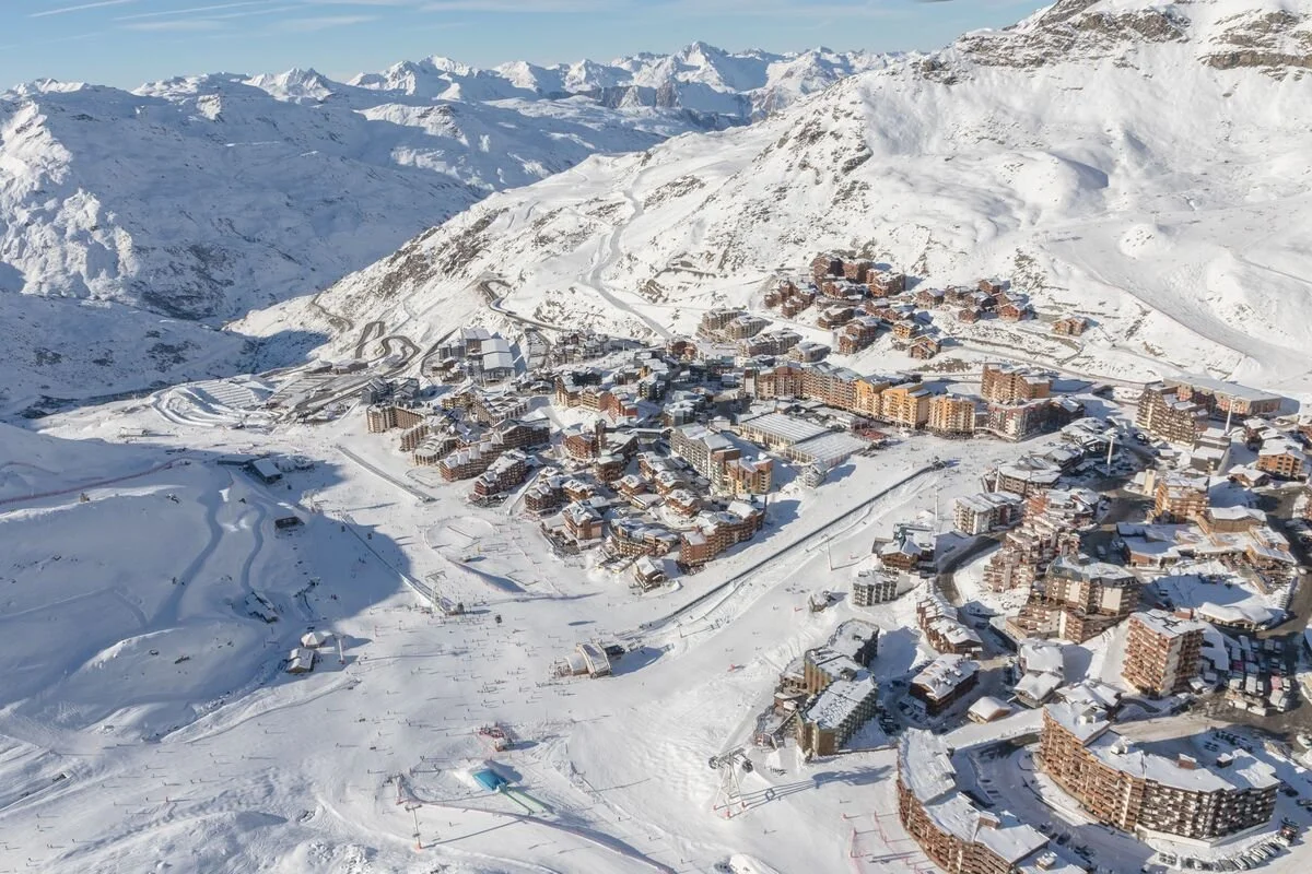 A snow-covered Val Thorens resort town with numerous buildings, ski slopes, and winding roads in a mountainous landscape.