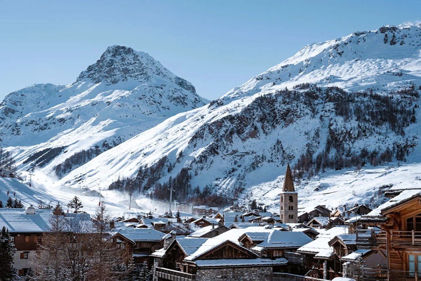 A snowy Val d'Isere mountain village with wooden houses, a church with a tall steeple, and snow-covered mountains in the background.