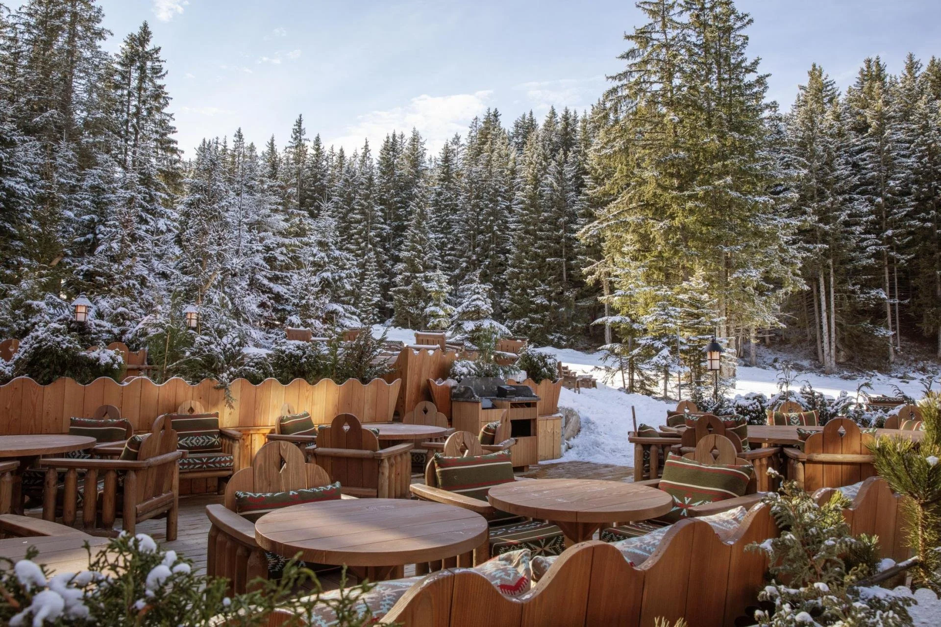 Outdoor patio area with wooden tables and chairs, surrounded by a snow-covered forest of tall pine trees under a partly cloudy sky, with decorative lanterns on the fence.