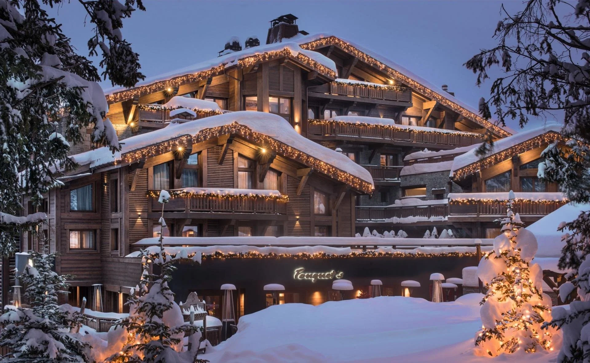 A multi-story wooden lodge or hotel decorated with string lights along the eaves, surrounded by snow-covered trees and ground, during the evening or nighttime.
