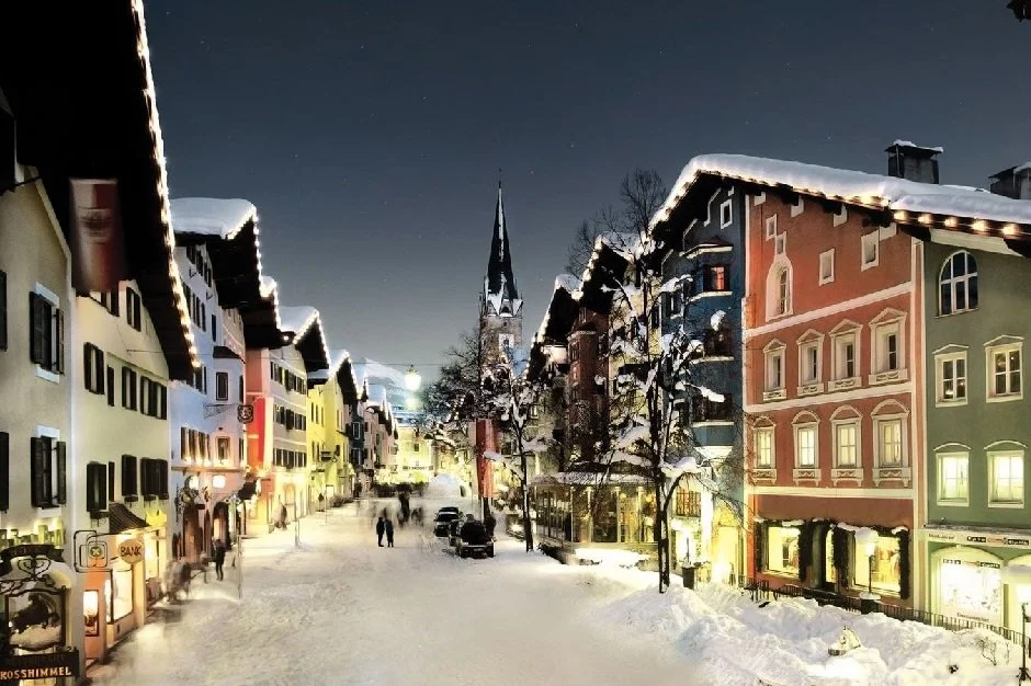 Snow-covered street in Kitzbuhel lined with colorful traditional European buildings and a church steeple in the background, illuminated at night.